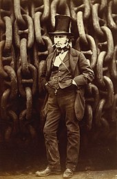 A 19th-century man wearing jacket trousers and waistcoat, hands in pockets, cigar in mouth, wearing a tall stovepipe top hat, standing in front of giant iron chains on a drum.