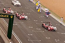 Four racing cars being driven on a motor racing circuit with people to their left waving flags and applauding them in congratulations.