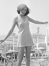 A dark-haired woman wearing a short dress, standing on a terrace overlooking the sea