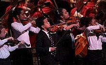 five children dressed in black and white play violins standing in front of seated adults in orange jackets