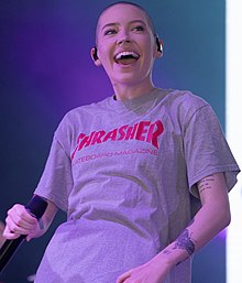 Porträtfoto einer lachenden Bishop Briggs. Sie trägt ein graues T-Shirt mit der roten Aufschrift Thrasher Skateboard Magazine, an beiden Armen kleine schwarze Tattoos, unter anderem einen Schmetterling, ein Herz sowie ein kurzes englischsprachiges Gedicht, und an beiden Ohren je zwei kleine, goldene Ohrringe. In der rechten Hand hält sie ein Mikrofon, in ihren Ohren steckt je ein kleiner, schwarzer Mikrofon-Stöpsel. Ihre Haare trägt sie als sehr kurz geschorene Stoppelfrisur.