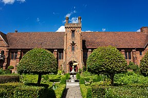 A Tudor brick building with a grand central entrance column. Green trees and neat topiary flank a sculpture in front of the Old Palace, Hatfield House