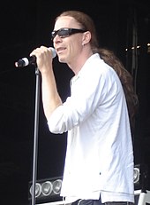 A picture of a man with long hair performing on stage with a microphone in his hand wearing sunglasses and a white top.