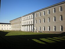 Long bâtiment blanc et rectangulaire, percé de nombreuses fenêtres, et haut de trois étage. La cour intérieure est gazonnée, on voit l'ombre d'une galerie avec colonnade, l'ensemble se dégage sur fon de ciel bleu.