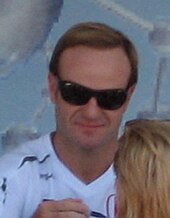 Photo de Barrichello, lunettes noires, signant un autographe à une femme blonde, vue de dos