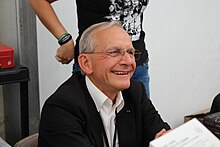 Un homme âgé aux cheveux gris est assis souriant.