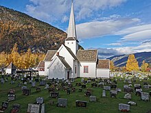 Foto einer kreuzförmigen weißen Holzkirche. Im Vordergrund stehen Grabsteine.