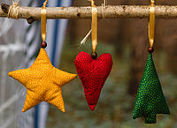 Holzzweig an dem 3 gefüllte Stoffbeutel in Form von Weihnachtssymbolen an goldenen Bändern hängen: links ein Stern in Gelb, in der Mitte ein Herz in Rot, rechts ein Tannenbaum in Grün. Der Hintergrund ist unscharf.