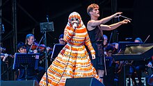 Image d'une femme dans une combinaison noir, blanc et orange chantant accompagnée d'un orchestre de cordes