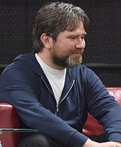 A man in his early 40s with brown hair and a brown beard looking to the right of the camera.