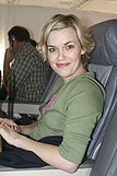 A woman sits in an airplane seat, smiling towards the camera. She has short blond hair.