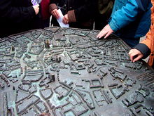 Blick von schräg oben auf ein bronzenes Relief, das modellhaft die Altstadt von Münster zeigt. Es ist in der Fußgängerzone der Münsteraner Altstadt etwa in Hüfthöhe horizontal auf einen Sockel montiert. Von den dicht dahinter stehenden Passanten sieht man nur die Hüften, einige betasten mit ihren Händen die Details des Modells. Dieses zeigt leicht stilisiert Straßen, Plätze, Gebäude und Baumgruppen; die Häuser sind etwa 2 cm hoch. Der Dom, die Lamberti- und die Liebfrauenkirche ragen mit mehreren Zentimetern Höhe deutlich heraus. Die Straßen sind in Brailleschrift mit ihren Namen gekennzeichnet. Am rechten Bildrand erkennt man Teile einer Widmung; dieser mehrzeilige Text ist sowohl in Brailleschrift als auch in erhabener Normalschrift ausgeführt.