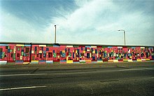 Frontale Farbfotografie von der Berliner Mauer mit einem Bild, das von der gegenüberliegenden Straßenseite aufgenommen wurde. Abstrakte Figuren sind in kleine, bunte Rechtecke gemalt, die auf rotem Untergrund sind. Über und unter dem Bild reihen sich bunte Querbalken aneinander.