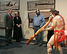 Pria Aborigin menampilkan Digeridoo indoors dengan 4 orang yang menonton, lukisan Aborigin dapat dilihat di tembok di belakangnya