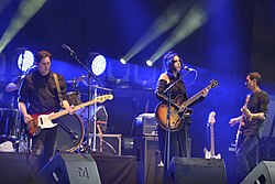 Chelsea Wolfe mit Gitarre vor einem Mikrofon auf der Bühne, links und rechts neben ihr zwei männliche Bandmitglieder; ebenfalls mit Gitarre und Bassgitarre; dahinter ein Schlagzeug zu erkennen
