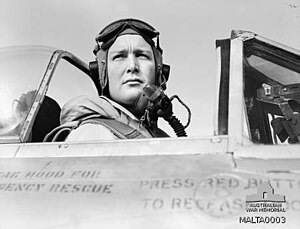 Black and white photo of a man wearing a flying helmet sitting in the cockpit of an aircraft