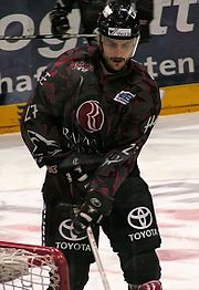 An ice hockey player standing directly in front of the camera. He is wearing a black helmet with a visor and a black and red uniform.