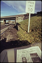 A sign next to a highway says "Speed Limit 50". A newspaper in the foreground has an article about the new speed limit.