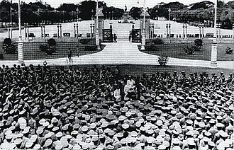 People gathering around a man standing on what is possibly a podium, reading a declaration, all of them wearing army- or related uniforms