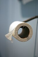 Toalettpappersrulle i hållare utan avrivningshjälpmedel, med pappret hängande framåt, över rullen, mot användaren