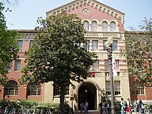 A building in a 19th-century style, with a four-story entrance in red brick and pale stone, framed with trees.