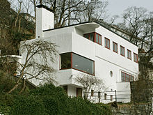 Una casa de estuco pequeña y con techo plano; diseñada con formas rectangulares simples, y construida sobre un cerro. Los marcos y los entramados de las ventanas son todos de madera.