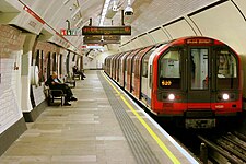 A deep level train stops to the right of a platform as some people (left) wait to board it.