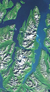 Satellitenbild, auf dem eine zerklüftete Landschaft zu sehen ist. Zentral befindet sich eine schmale, von Fjorden umgebene Halbinsel. Auf der Halbinsel befinden sich schneebedeckte Berge, an der Küste der Halbinsel ein grüner Streifen