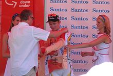 A cyclist is presented an orange jersey, and is in the process of donning it. A man wearing white is facing the cyclist and pulling the jersey's sleeves forward around him. Two women in white dresses flank the cyclist.