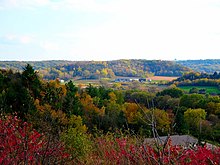 Landscape with low rolling hills, farm buildings in the middle distance surrounded by fields, and bright early autumn foliage