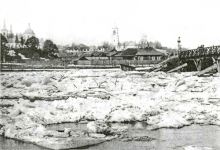 Photographie en noir et blanc de la rivière partiellement gelée, avec le village en arrière-plan.