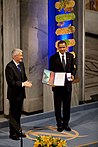 Two men standing on a stage. The man to the left is clapping his hands and looking towards the other man. The second man is smiling and showing two items to an audience not seen on the image. The items are a diploma which includes a painting and a box containing a gold medal. Behind them is a blue pillar clad in flowers.