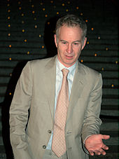 A man in a grey suit with a pink tie, with his hand out in front
