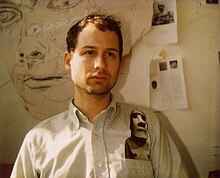 Young man in an art studio, in front of a large drawing and art clippings taped to the wall.