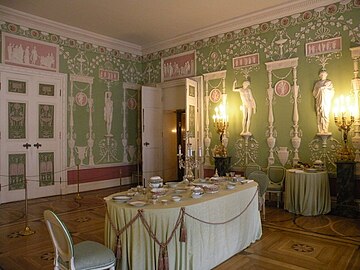 Farbige Eckansicht eines Innenraums mit einem gedeckten Tisch und Stühlen in der Mitte. An den hellgrünen Wänden sind viele weiße Ornamente und drei antike Frauenfiguren angebracht. Über den Figuren und über beiden Türen sind rosa Reliefs mit weißen Szenen zu sehen. An den Türflügeln sind grün-rosa Muster eingezeichnet. Vor der rechten Wand strahlen zwei Stehleuchten.