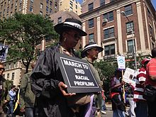 Demonstration. Im Bildvordergrund eine schwarze Frau mit gesenkten Blick. Sie trägt mit beiden Händen ein schwarzes Schild mit der Aufschrift: „MARCH TO END RACIAL PROFILING“