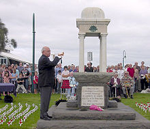 A balding man wearing a suit and playin a bugle, while staundin in front o a croud o ither fowk an a stane monument.