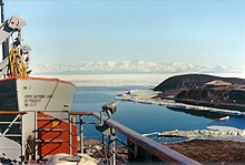 In left foreground are the upper works of a modern ship. To the right, on a low headland fringed with sea ice, is a square hut with a pitched roof. In the distance is a range of icy mountains