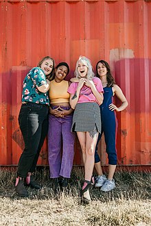 Four women standing in front of an orange-red wall, they are all smiling or laughing