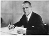 A photographic portrait of a man wearing a suit seated at a desk with a pen in his hand