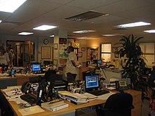 Photo prise à l'intérieur d'un bâtiment présentant une série de bureaux avec des ordinateurs et du papier au sein d'un open-space. Des personnes de l'équipe technique sont présentes à l'arrière-plan.