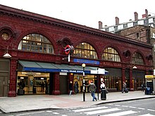 A red glazed terracotta building. The first storey above ground features four wide, storey-height semi-circular windows with smaller circular windows between above which is a dentil cornice. Below the two right-most windows, the station name, "Russell Square Station", is displayed in gold lettering moulded into the terracotta panels. A blue tiled panel above the entrance says "Underground".