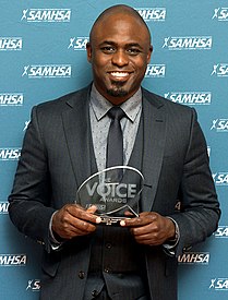 Wayne Brady steht vor einer blauen Wand mit der weißen Aufschrift SAMHSA. Er trägt ein dunkelblaues Jackett, ein weißes Hemd mit kleinen schwarzen Punkten und eine schwarze Krawatte mit kleinen weißen Punkten. In den Händen hält er eine ovale, auf einem kleinen Sockel stehende Auszeichnung mit der Aufschrift The Voice Awards.