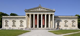 Frontale Farbfotografie eines weißen Gebäudes mit Säuleneingang und einem Giebelfeld, das eine antike Szene darstellt. Auf dem Dach stehen drei Ornamente und hinter den Säulen hängen zwei vertikale Banner mit der Aufschrift „Die unsterblichen Götter Griechenlands“. An beiden Seiten stehen jeweils drei weiße Statuen in Nischen, die von Giebelfeldern überdacht sind. An den vorderen Stufen sitzen Menschen und ein breiter Mittelweg mit Rasenflächen führt zum Gebäude.