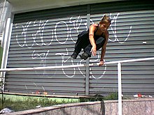 A white woman in black clothes supports herself on her left arm over a white rail. In the background a roller shutter has graffiti.