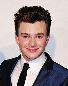 Photo de l'acteur Chris Colfer, alors jeune homme, souriant : il est rasé de près, a des cheveux courts coiffés avec du gel, et est habillé en costume-cravate.