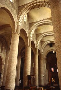 A church interior of yellow stone with arches of alternating red and cream crossing the nave to support an unusual vaulting system.