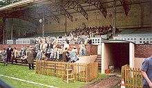 Scène de tournage dans les tribunes d'un stade de football, en couleur.