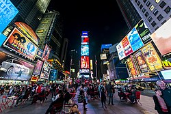 many pedestrians, drivers, and seated people active at night and lit by streetlights, headlights, and billboards