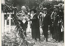 Photographie en noir et blanc d'un homme, accompagné d'une femme qui lui tend un bouquet de fleurs, et une rangée d'hommes.